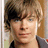 A pixelated image of a person with light brown hair and blue eyes, wearing a dark jacket over a black shirt against a blurred background.