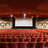 Static pixelated image of a theater interior with a large screen, dark frame, and patterned red-brown seating area.