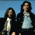 Two figures with long dark hair stand side-by-side against a blue background, wearing dark outer garments over light-colored shirts.