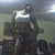 A person in a dark sleeveless top stands in a dimly lit room with a -patterned chair and a light fixture visible in the background
