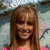 A person with long brown hair wears a pink crop top and white skirt, smiling with a light blue background.