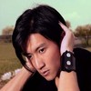 A person with dark hair holds both hands near their head, wearing a black shirt and a black wristwatch with white buttons, against a blurred outdoor background.