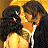 Two figures in close physical contact with dark hair, one in a light-colored dress, the other in dark clothing with a white stripe, against a warm-toned background.