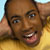 Pixelated close-up of a person with dark skin wearing a yellow shirt, smiling with mouth open showing teeth, hand raised near head, light blue and yellow background.