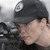 A pixelated close-up of a person wearing a dark cap with a circular white emblem, holding a hand near their face against a blurred background.