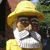 Pixelated image of a figure wearing a yellow wide-brimmed hat, dark glasses, and a white beard, with a yellow garment visible against a blurred background.