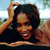 A person with dark hair wearing a dark top, raising one arm above their head against a blurred outdoor background.