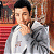 A pixelated image of a person with short dark hair wearing a light gray jacket over a white shirt, standing against a blurred background with red and brown tones.