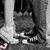 Black-and-white pixelated image showing two people sitting closely together on a bench, with one person's legs and feet visible in the foreground.