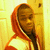 Pixelated image of a person with dark skin wearing an orange hooded garment over a white shirt, standing against a yellow background.