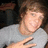 Pixelated image of a person with light brown hair wearing a light gray shirt, making a hand gesture with fingers extended against a dark background.