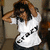 A person in a white shirt with black text adjusts their hair against a neutral background.