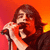 A pixelated image of a person with long brown hair singing into a microphone against a gradient background of red and orange hues.