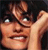 A pixelated close-up of a person with dark hair, wearing glasses, and holding a hand near their face, set against a dark background.