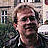 Pixelated portrait of a person with light brown hair wearing dark clothing against a blurred background with indistinct shapes and colors.