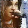 A close-up of a person with light brown hair holding a microphone near their mouth, wearing a white garment with an orange collar.