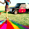 A woman in a blue top and patterned skirt runs on grass with a black car and rainbow reflection in the background.