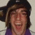 A person with brown hair wearing a black cap and purple shirt, smiling broadly across three