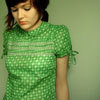 A person in a green checkered dress with short sleeves and a tied detail on the right sleeve, standing against a light green background.