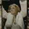 A woman in a white jacket with curly hair performs animated gestures against a dark background.