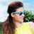 A person with long brown hair in a ponytail wears a yellow sleeveless top and light blue sunglasses, standing outdoors with green trees and a blue sky background.
