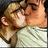 Two figures in close physical proximity, one with light brown hair and the other with darker hair, sharing a kiss against a dark background.