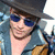 A person wearing a dark wide-brimmed hat, blue sunglasses, and a light blue jacket over a brown shirt, with a neutral expression.