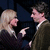 A woman with long blonde hair points her right index finger at a man in a brown suit, both smiling against a dark blue background.