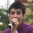 A person with dark hair wearing a purple shirt holds a microphone near their mouth against an outdoor background.