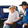 Two people sit in a vehicle; one wears a light cowboy hat and white sweater, the other a dark cap and blue jacket.