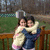 Two people embrace on a wooden deck with green grass and trees in the background, captured in a static image with no visible motion.