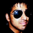 Pixelated close-up of a person's face with light brown hair, dark sunglasses, and a neutral expression against a black background.