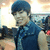A person with short dark hair wearing a dark blue sleeveless shirt with light blue patterns, smiling and holding a camera in a bright indoor setting.