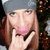 Pixelated image of a person with long brown hair wearing a gray beanie, holding a black object near their face with a pink mark on the cheek.