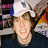 Pixelated static image of a person with light skin and brown hair wearing dark clothing, set against an indistinct background with blurred colors including pink, blue, yellow, and red.