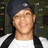 Pixelated close-up of a person with light skin wearing a dark cap and white shirt, set against a blurred background.