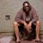 A pixelated static image shows a person with long hair and a beard wearing a light-colored shirt and dark shorts, seated with legs apart against a plain wall background.