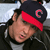Static pixelated image of a person wearing a dark cap with red brim and circular logo, visible ear, and partial clothing in grayscale tones.