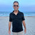 Pixelated static image of a person standing on a beach wearing a dark polo shirt and light pants against a blue sky and water background.
