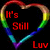 A heart-shaped rainbow outline with red text reading "It's Still Luv" on a black background, next to a rainbow arch over two white clouds.