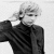 Black-and-white pixelated static image of a person with light-colored hair wearing dark clothing, viewed from the shoulders up against a plain background.