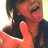 A person with dark hair and a black top holds up two orange hands in front of their face, making a playful gesture.