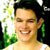 Pixelated static image of a person with dark hair and light skin, smiling broadly in an outdoor setting with green foliage on the left side.