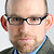 Pixelated close-up of a person wearing dark-framed glasses, light blue shirt, and dark jacket against a light background.