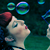 Pixelated image of a light-skinned figure with blue eyes and red lips looking upward at floating translucent bubbles against a dark teal background.