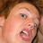 Pixelated close-up of a person's face with light skin, brown hair, and an open mouth showing teeth, set against a blurred background.