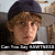 Pixelated image of a person with light brown hair wearing a dark blue cap, set against a blurred background with visible text at the bottom.