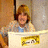 Pixelated image of a person with light brown hair wearing a blue top, seated behind a white counter with a yellow sign displaying partial text.