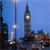 Pixelated cityscape featuring a tall clock tower with a visible clock face, adjacent to a slender spire, under a clear blue sky with scattered buildings below.