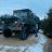Pixelated static image shows dark vehicle on light ground under blue sky with trees.